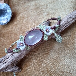 May include: A handmade macrame bracelet with a large, oval rose quartz stone. The bracelet uses brown and green macrame cord, with delicate floral accents in light blue and gold. The bracelet is displayed on a piece of driftwood.