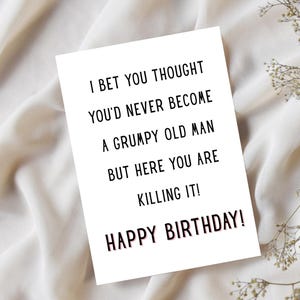 May include: A white greeting card with black text that reads "I BET YOU THOUGHT YOU'D NEVER BECOME A GRUMPY OLD MAN BUT HERE YOU ARE KILLING IT! HAPPY BIRTHDAY!". The card is on a white fabric background.