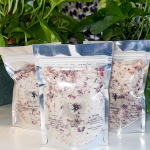 Może przedstawiać: Trzy srebrne saszetki wypełnione białą mieszanką soli do kąpieli z suszonymi płatkami róż. Saszetki mają czarno-białe logo z tekstem "EVEN A LITTLE... Rejuvenate Bath Salt".