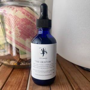 Könnte beinhalten: Eine dunkelblaue Glasflasche mit schwarzer Pipette, beschriftet mit "LIFE'S AMULET" und "THE DEEP OIL". Die Flasche steht auf einer Holzoberfläche, im Hintergrund ein Glasgefäß mit Papiergeld.