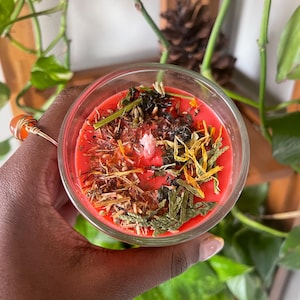 May include: A red candle with dried herbs and flowers embedded in the wax. The candle is in a clear glass jar.