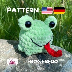 May include: A light green crocheted frog with black eyes and a red tongue. The frog is labeled "FROG FREDO" and the word "PATTERN" is above the frog. The frog is sitting on a rock with green foliage in the background.