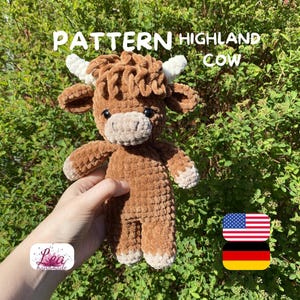 Könnte beinhalten: Gehäkeltes Highland-Kuh-Spielzeug in Braun- und Beigetönen mit weißen Hörnern und einem flauschigen Kopf. Der Text "PATTERN HIGHLAND COW" ist im Bild enthalten. Das Spielzeug wird vor einem Hintergrund aus grünem Laub gehalten.