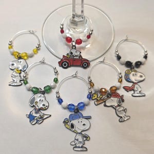 Peut inclure: Ensemble de six marque-verres sur le thème de Snoopy. Chaque breloque est composée d'un anneau argenté orné de perles colorées et d'un charm représentant Snoopy. Snoopy est représenté dans diverses poses, dont au volant d'une voiture rouge et jouant au baseball.