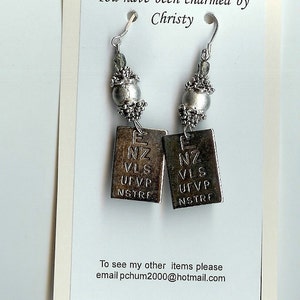 Pode incluir: Brincos prateados com pingentes retangulares, cada um gravado com letras e números. Os brincos têm um detalhe de contas e são exibidos em um cartão que diz "You have been charmed by Christy."