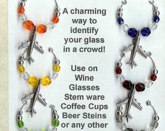 May include: Six silver-toned airplane wine glass charms arranged on a display card. Each circular charm features a metal airplane pendant and faceted beads in orange, yellow, green, purple, red, and grey. The card includes text about glass identification.