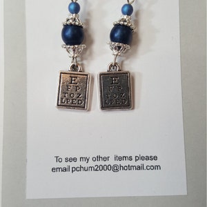 Pode incluir: Brincos pendurados com ganchos prateados. Cada brinco apresenta uma conta azul escura, um elemento decorativo prateado e um pequeno pingente retangular com as letras "E FP TOZ LPED". O texto "You have been charmed by Christy" também é visível.