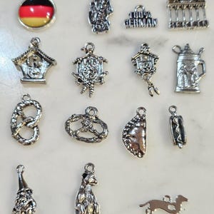 May include: A collection of silver-toned charms with German themes. Charms include a German flag, cuckoo clocks, pretzels, a beer stein, a gnome, a bear, a dachshund, and the text "100% GERMAN". These charms are ideal for jewelry making or crafting.