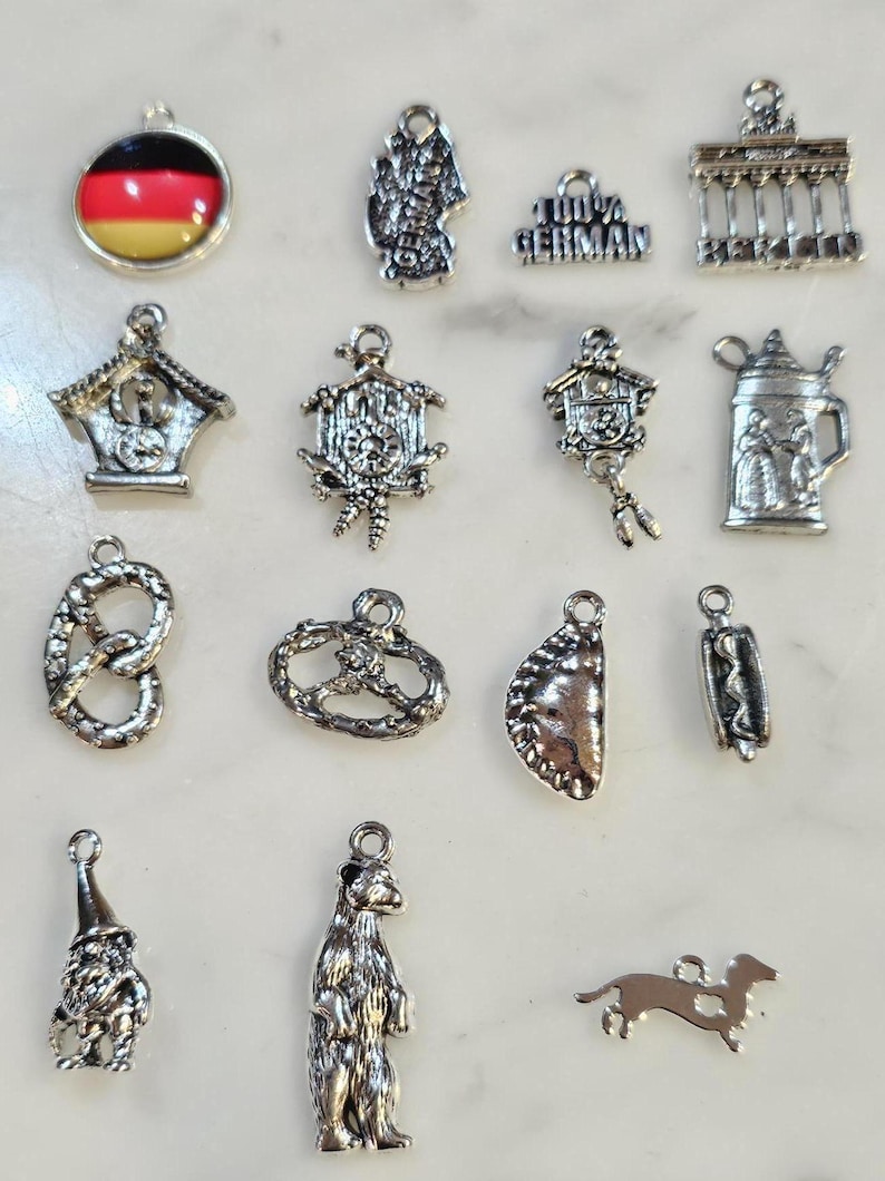May include: A collection of silver-toned charms with German themes. Charms include a German flag, cuckoo clocks, pretzels, a beer stein, a gnome, a bear, a dachshund, and the text "100% GERMAN". These charms are ideal for jewelry making or crafting.