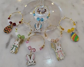 May include: A selection of Easter wine charms. Each charm has a gold-tone hoop with colourful beads and a dangling Easter-themed charm, including eggs and bunnies. The charms are designed to be attached to wine glasses.