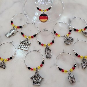 May include: A collection of wine glass charms with silver hoops and decorative charms. The charms feature red, yellow, and black beads, and miniature German-themed pendants, including a beer stein, Brandenburg Gate, and a pretzel.