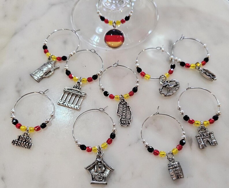 May include: A collection of wine glass charms with silver hoops and decorative charms. The charms feature red, yellow, and black beads, and miniature German-themed pendants, including a beer stein, Brandenburg Gate, and a pretzel.