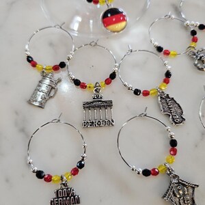 May include: Wine glass charms with German-themed pendants. Each charm features a silver hoop with black, red, and yellow beads. Charms include a German flag, a beer stein, the Brandenburg Gate, and the text "100% GERMAN".
