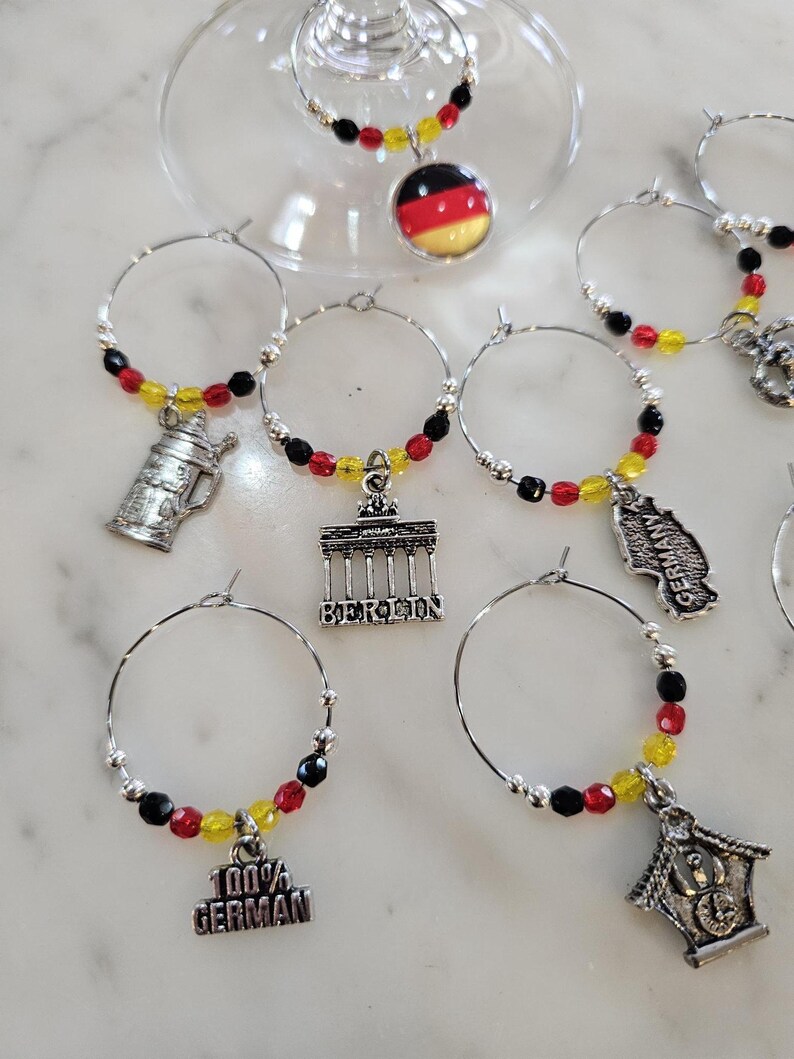 May include: Wine glass charms with German-themed pendants. Each charm features a silver hoop with black, red, and yellow beads. Charms include a German flag, a beer stein, the Brandenburg Gate, and the text "100% GERMAN".