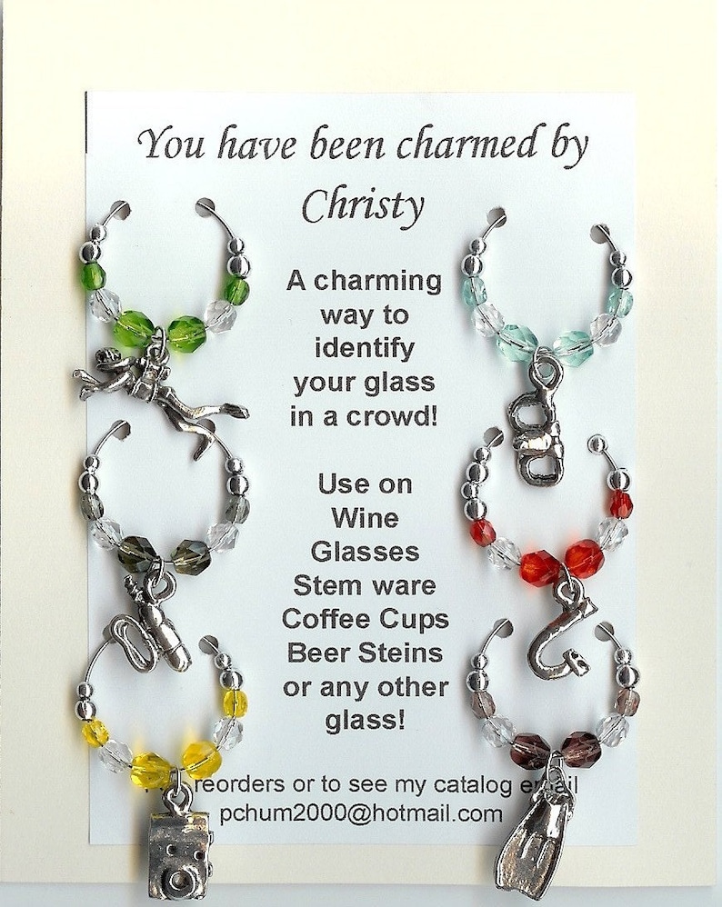 May include: Wine glass charms with various charms and colorful beads. The charms include a diver, a number 8, a saxophone, a camera, a letter G, and a diving fin. Text on the card reads "You have been charmed by Christy."