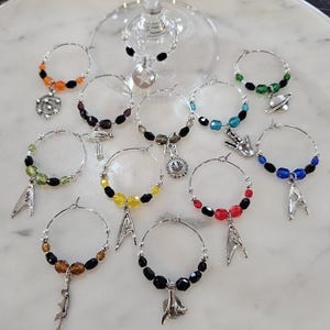 May include: A collection of wine glass charms with silver hoops, colorful beads, and themed charms. The charms feature various colors of beads, including orange, green, blue, and black. The charms have silver pendants with space-themed designs.