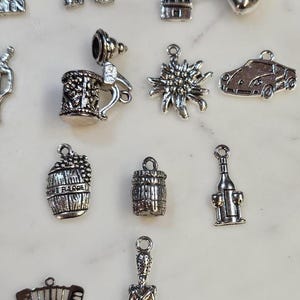 May include: A collection of silver-colored charms, including a beer stein, a windmill, a car, a wine barrel, and an accordion. The charms are detailed and have a vintage aesthetic, suitable for jewelry making or crafting.