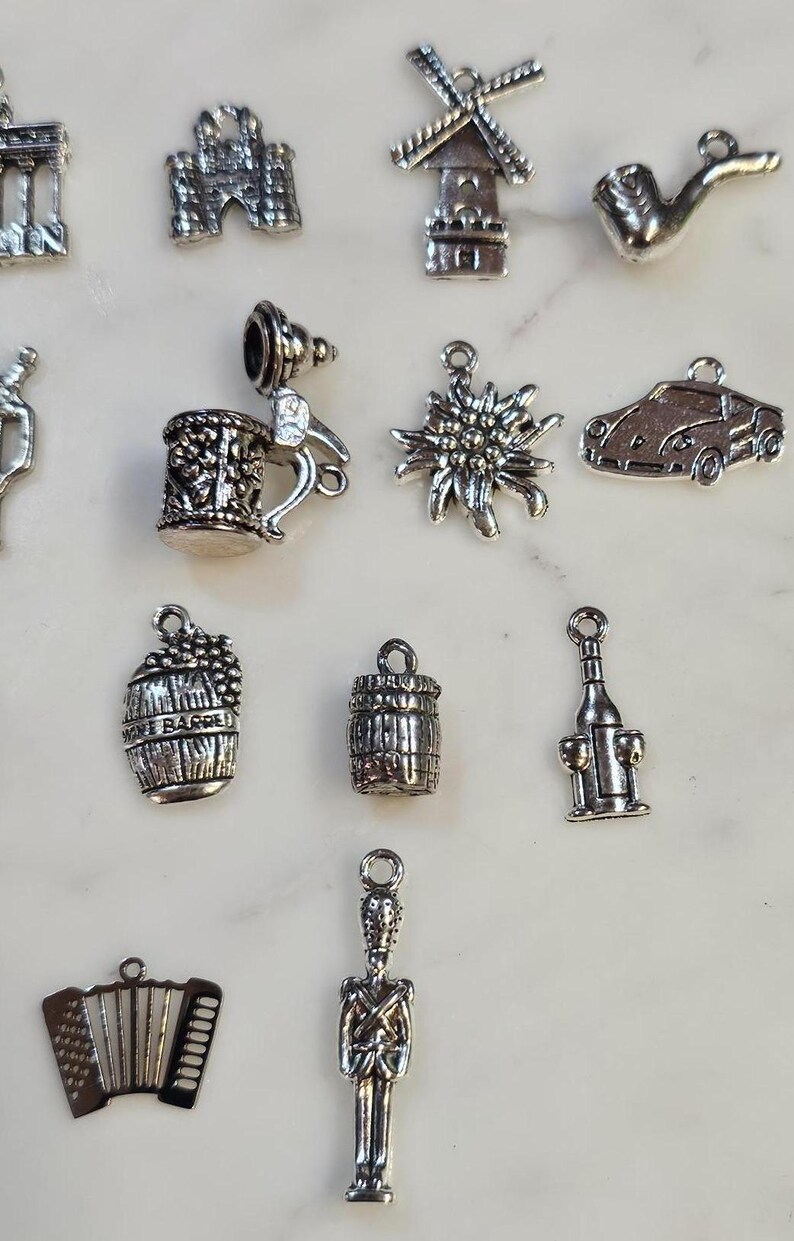 May include: A collection of silver-colored charms, including a beer stein, a windmill, a car, a wine barrel, and an accordion. The charms are detailed and have a vintage aesthetic, suitable for jewelry making or crafting.