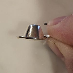May include: A small, shiny, silver-colored metal object, shaped like a miniature hat. The object is held between two fingers. The number "28" is visible in the background.