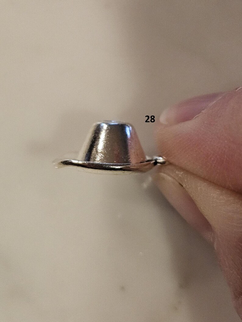 May include: A small, shiny, silver-colored metal object, shaped like a miniature hat. The object is held between two fingers. The number "28" is visible in the background.