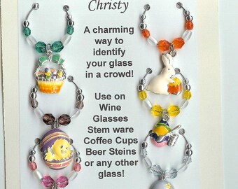 May include: A set of four circular metal wine glass charms on a display card with the text You have been charmed by Christy. The charms feature colourful enamel pendants including an Easter basket, a white rabbit, a chick in an egg, and a floral egg.