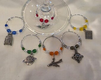 May include: A collection of wine charms with travel-themed pendants. Each charm features a silver ring adorned with colorful beads and a unique charm, including a suitcase, airplane, and globe. The charms are designed to hang on a wine glass.