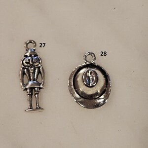 May include: Two silver-toned charms. One charm is a detailed miniature of a nutcracker soldier. The other charm is a cowboy hat with a detailed brim and crown. Both charms have a loop for attaching to a necklace or bracelet.