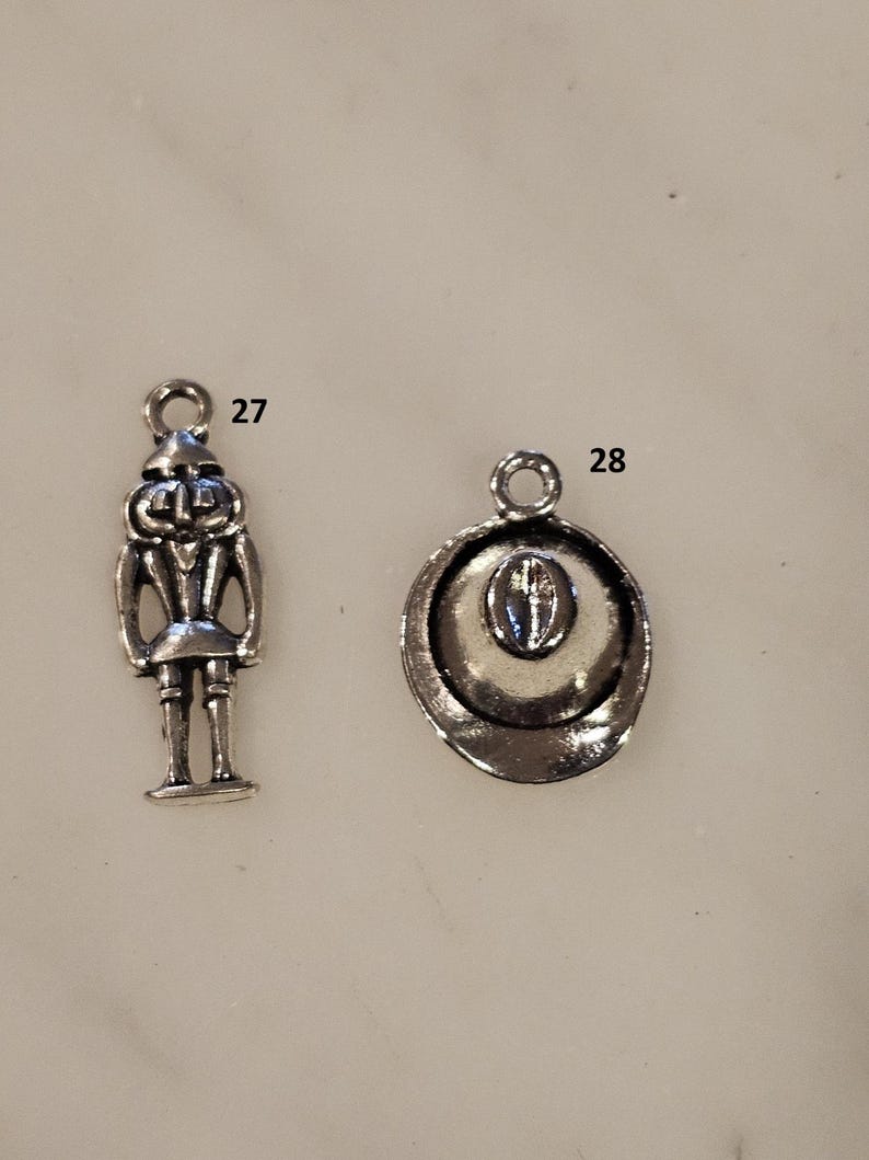 May include: Two silver-toned charms. One charm is a detailed miniature of a nutcracker soldier. The other charm is a cowboy hat with a detailed brim and crown. Both charms have a loop for attaching to a necklace or bracelet.