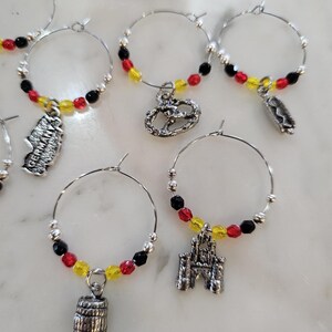 May include: Wine charms with German-themed designs. Each charm features a silver hoop with red, yellow, and black beads, and a German-themed charm. Charms include a pretzel, a barrel, a castle, and a map of Germany.