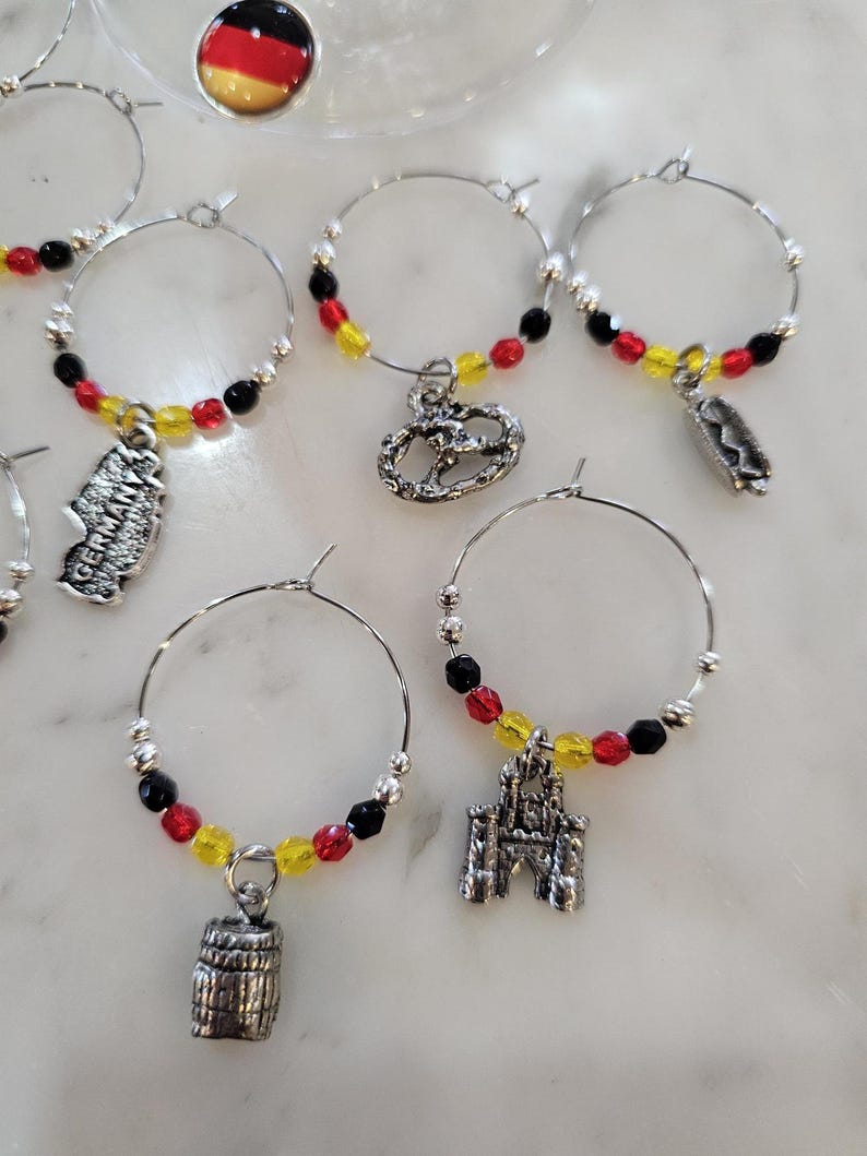 May include: Wine charms with German-themed designs. Each charm features a silver hoop with red, yellow, and black beads, and a German-themed charm. Charms include a pretzel, a barrel, a castle, and a map of Germany.