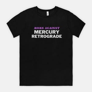 May include: Black t-shirt with the text "MOMS AGAINST MERCURY RETROGRADE" in white and purple lettering. The shirt is a crew neck style and appears to be made of cotton or a similar fabric. The text is centered on the front of the shirt.