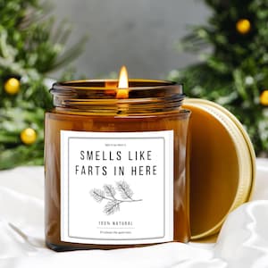 May include: A lit brown glass jar candle with a white label that reads "SMELLS LIKE FARTS IN HERE". The candle has a gold lid and a pine branch illustration. The text "100% NATURAL" is also on the label.