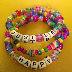 May include: Three colourful beaded bracelets. One spells "JUST BE", another "HAPPY". The bracelets feature a variety of beads in pink, blue, green, and yellow. The bracelets are on a yellow background.