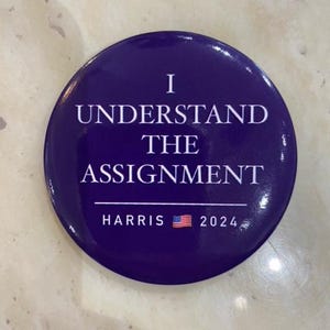 May include: A circular, deep purple badge with white text reading "I UNDERSTAND THE ASSIGNMENT". Below, it says "HARRIS 2024" with a small American flag graphic.