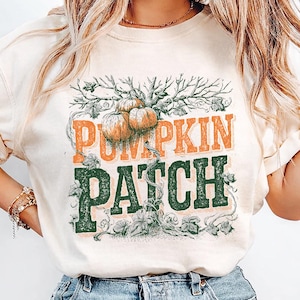 Könnte beinhalten: Ein cremefarbenes Kurzarm-T-Shirt mit einem Grafikdruck von orangefarbenen Kürbissen an Ranken sowie dem Text PUMPKIN in Orange und PATCH in Dunkelgrün. Das Design zeigt detaillierte Linienzeichnungen von Zweigen und Blättern.