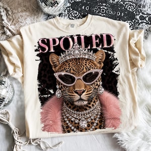 May include: Cream-colored t-shirt with a graphic of a leopard wearing sunglasses, a crown, and jewelry. The word "SPOILED" is printed above the leopard in pink letters. The leopard has a pink fur collar.