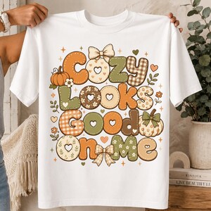 May include: A white t-shirt featuring a graphic print with the text "Cozy Looks Good On Me" in a stylized, multi-colored font. The design includes illustrations of pumpkins, bows, and heart shapes in shades of orange, green, and beige.