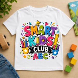 May include: White t-shirt with a colorful graphic that reads "SMART KIDS CLUB ABC". The design includes pencils, a ruler, stars, and a lightbulb. The shirt is displayed on a wooden surface with toys and a book.