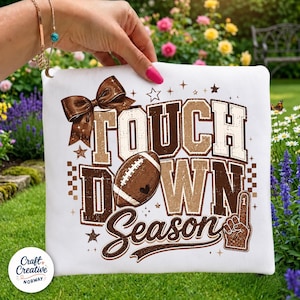 May include: A white fabric item featuring a brown graphic print with the text "TOUCH DOWN SEASON". The design includes a large brown bow, a football, a foam finger, and small star accents. The lettering has a distressed, textured appearance.