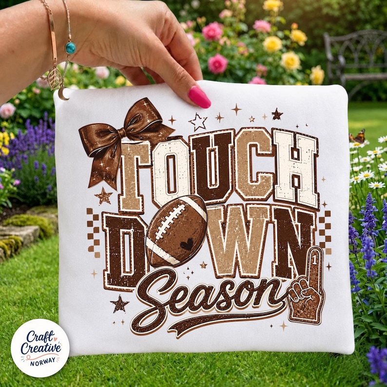 May include: A white fabric item featuring a brown graphic print with the text "TOUCH DOWN SEASON". The design includes a large brown bow, a football, a foam finger, and small star accents. The lettering has a distressed, textured appearance.