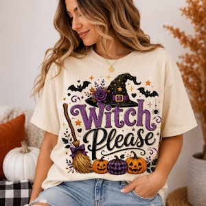 May include: A cream-colored t-shirt featuring a graphic print with the text "Witch Please" in purple and black. The design includes a black witch hat with a purple band, a broomstick, three pumpkins, bats, stars, and a purple flower.