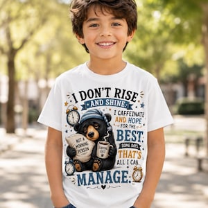 K&ouml;nnte beinhalten: Ein wei&szlig;es Kurzarm-T-Shirt mit einer Grafik eines schl&auml;frigen schwarzen B&auml;ren mit blauer Schlafm&uuml;tze, der ein Kissen mit der Aufschrift 'NOT A MORNING PERSON' und eine Tasse mit 'SURVIVAL JUICE' h&auml;lt, umgeben von Weckern und dem Text 'I DON'T RISE AND SHINE'.