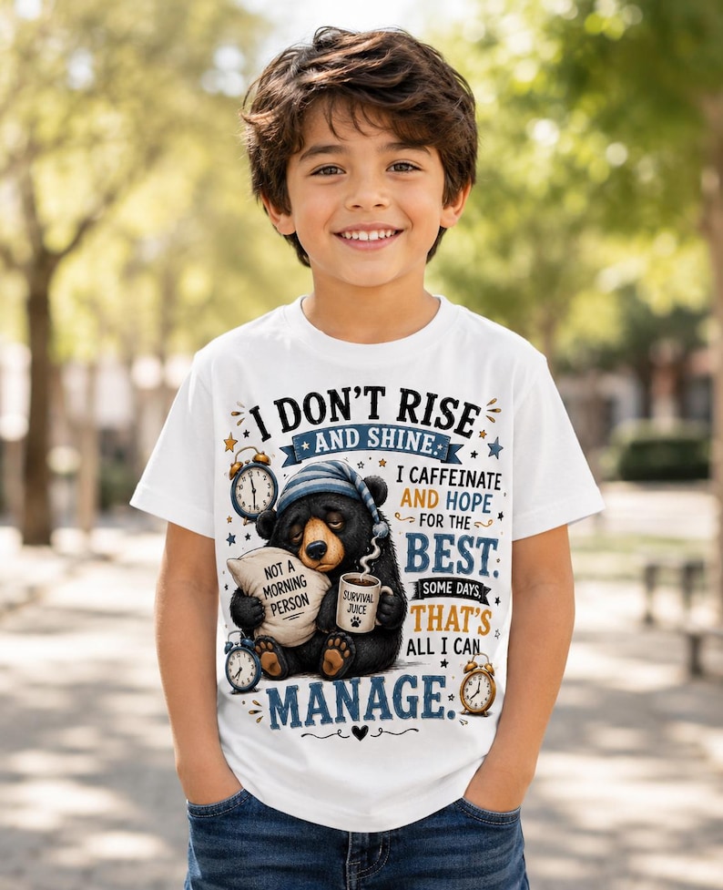 K&ouml;nnte beinhalten: Ein wei&szlig;es Kurzarm-T-Shirt mit einer Grafik eines schl&auml;frigen schwarzen B&auml;ren mit blauer Schlafm&uuml;tze, der ein Kissen mit der Aufschrift 'NOT A MORNING PERSON' und eine Tasse mit 'SURVIVAL JUICE' h&auml;lt, umgeben von Weckern und dem Text 'I DON'T RISE AND SHINE'.