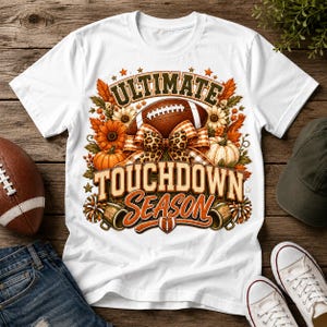 Op de afbeelding: Een wit t-shirt met een grafische print met de tekst "ULTIMATE TOUCHDOWN SEASON". Het ontwerp bevat een bruine football, oranje pompoenen, zonnebloemen, een strik met luipaardprint en twee pompons in een herfstachtig arrangement.