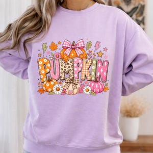 May include: A light purple crewneck sweatshirt featuring a graphic print with the word PUMPKIN in large, patterned letters. The design includes orange and pink pumpkins, a pink plaid bow, autumn leaves, flowers, stars, and lightning bolts.