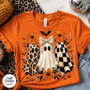 May include: An orange t-shirt featuring a graphic of three ghosts. The left ghost has a leopard print pattern, the center ghost wears a large pearl-embellished bow, and the right ghost has a black and white checkerboard pattern, surrounded by orange flowers and bats.