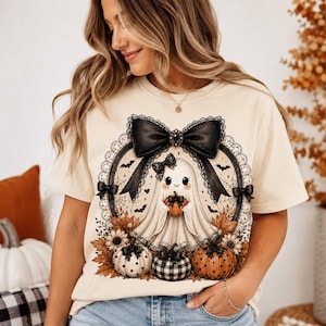 May include: A cream-colored short-sleeve t-shirt featuring a graphic of a white ghost holding a small orange pumpkin. The ghost is framed by a black lace border, a large black bow, and surrounded by autumn leaves, sunflowers, and pumpkins.