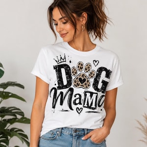 May include: White t-shirt with the text "DOG Mama" in black, featuring a paw print with a leopard print design and a heart. The design also includes a crown and hearts. The shirt is worn by a person.