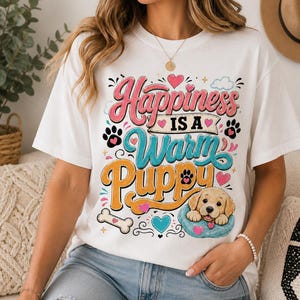 May include: A white short-sleeved t-shirt featuring a colorful graphic print with the text "Happiness IS A Warm Puppy". The design includes a golden puppy resting in a blue bed, paw prints, hearts, and a bone, rendered in pink, blue, and orange.