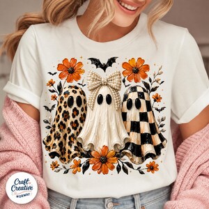 May include: A white short-sleeve t-shirt featuring a graphic of three ghosts. The left ghost has a leopard print, the center ghost wears a pearl-embellished bow, and the right ghost has a black and white checkerboard pattern, surrounded by orange flowers and bats.